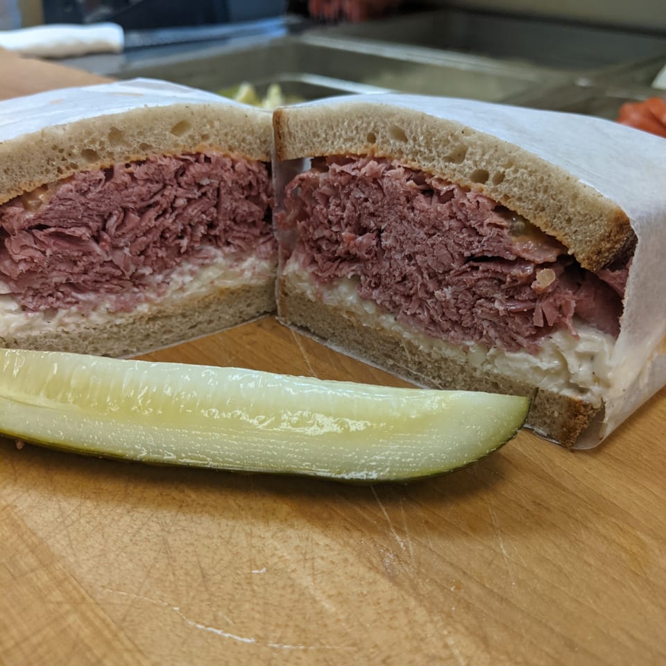 A deli sandwich, cut in half, with a pickle spear on a wooden cutting board.