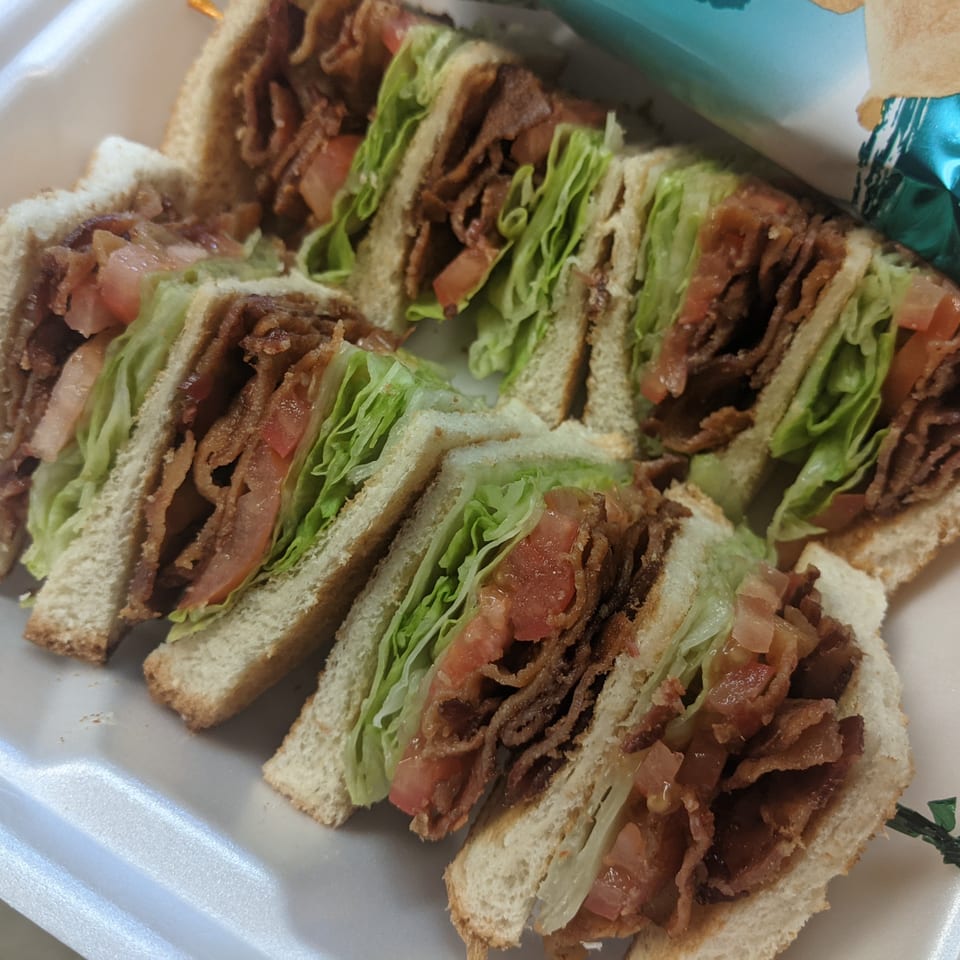 Several club sandwiches with bacon, lettuce, and tomato, cut into triangles in a styrofoam container.
